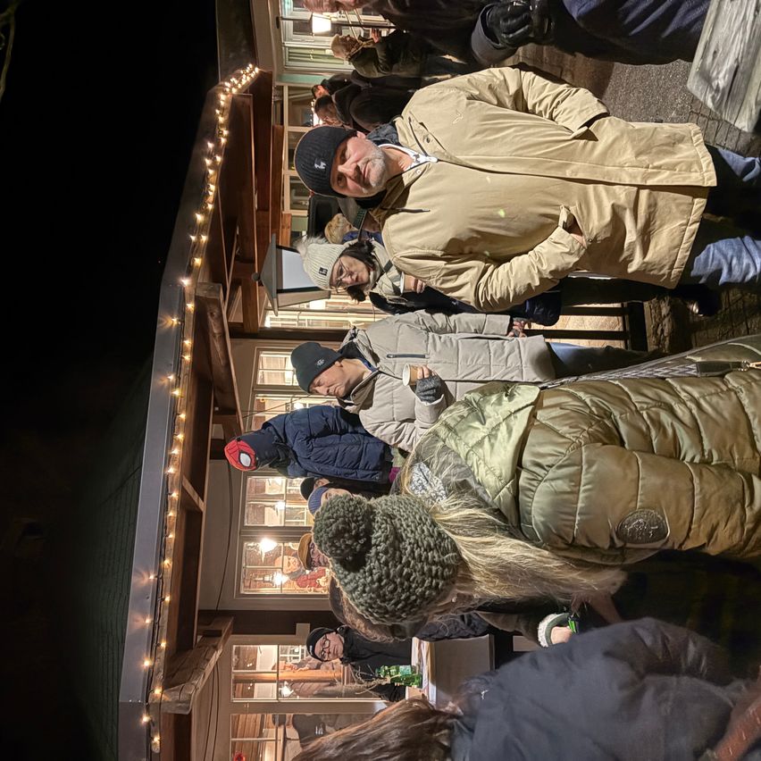 Eine Gruppe von Menschen in Winterkleidung, Mützen und Handschuhen steht in einer Schlange, wahrscheinlich warten sie darauf, einen Ort zu betreten. Es gibt Lichter und Fenster am Gebäude.