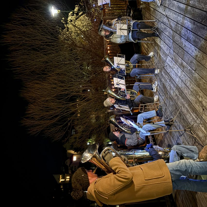 Ein nächtliches Open-Air-Konzert mit einem Brass-Ensemble, bei dem Musiker auf einer Holzplattform unter einer Straßenlaterne Posaunen spielen.