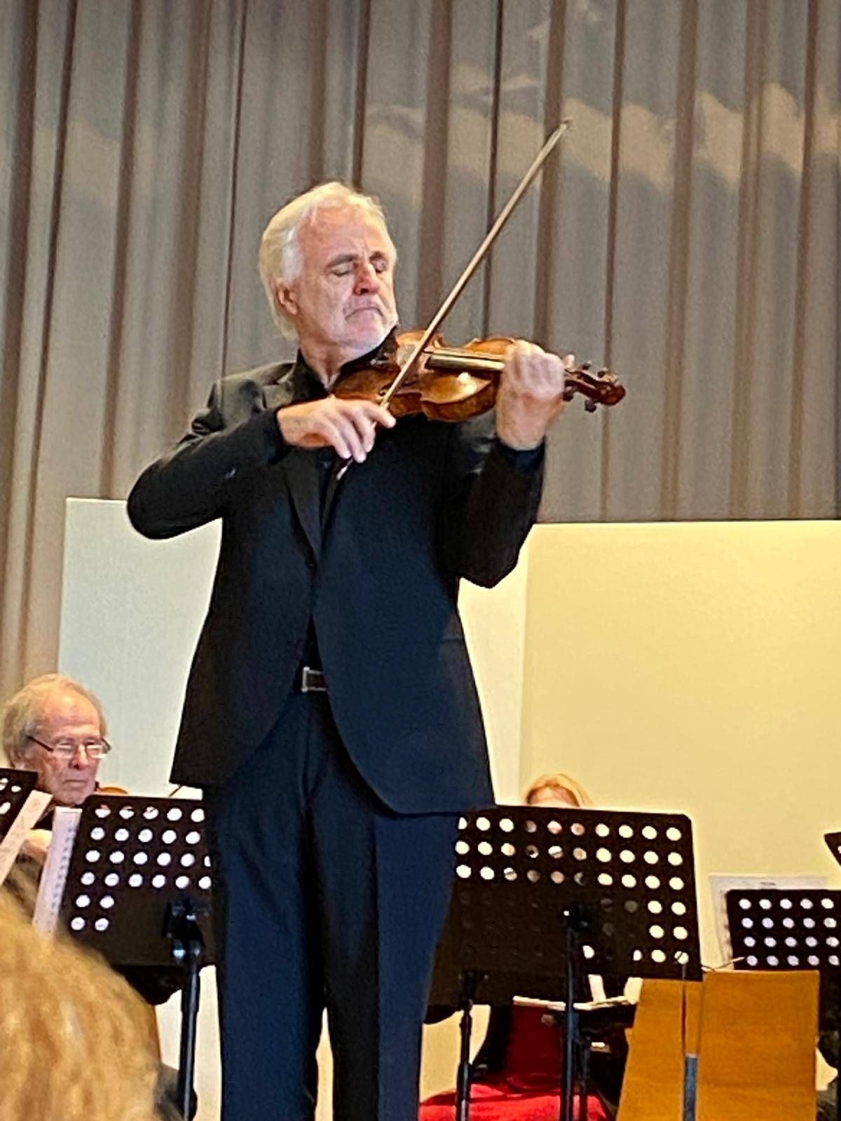 Ein älterer Mann spielt Geige auf der Bühne in einem schwarzen Anzug, mit einem anderen Mann, der hinter ihm spielt, und einem Vorhang als Hintergrund.