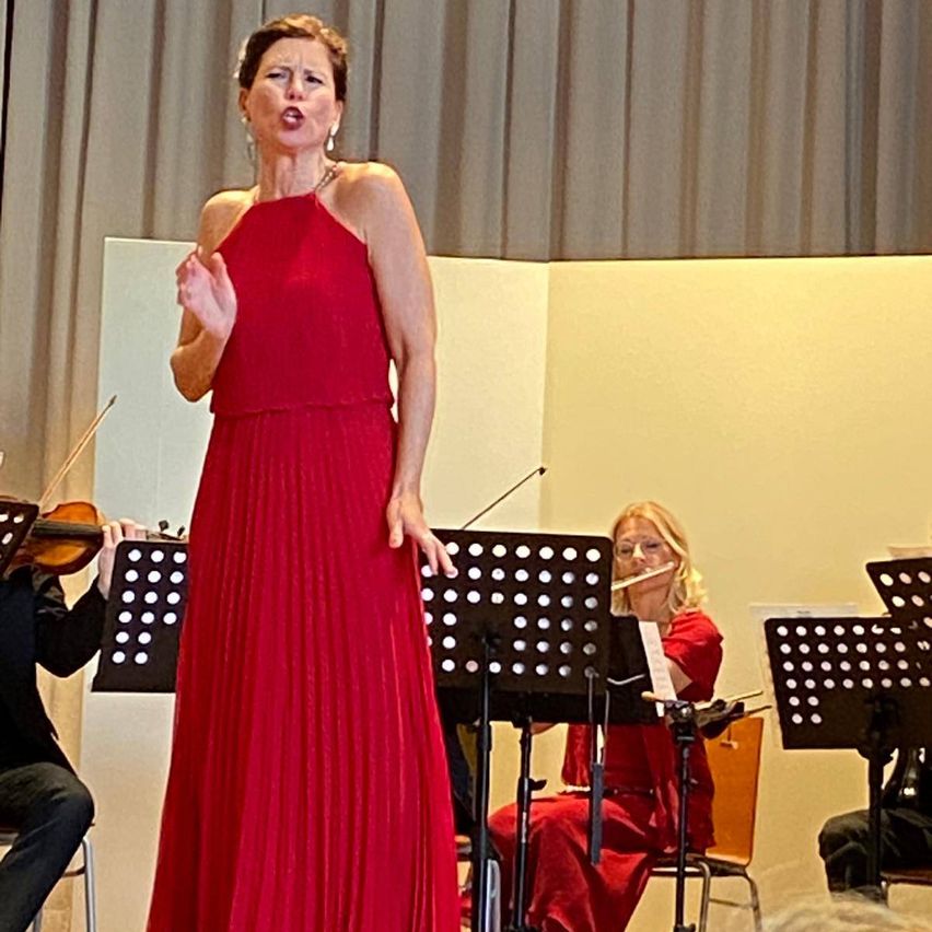 Eine Frau in einem roten Kleid singt auf der Bühne, während ein Violinist und eine Flötistin hinter ihr spielen. Die Musiker sitzen auf Stühlen mit Notenständern vor ihnen.