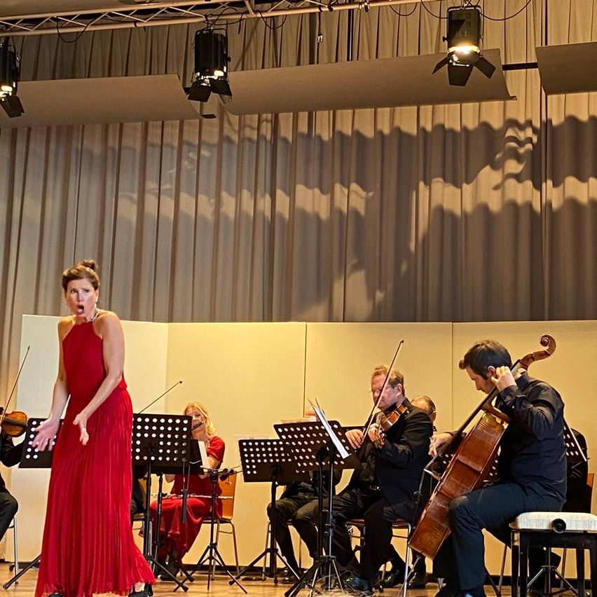 Eine Frau in einem roten Kleid tritt auf der Bühne auf, während ein Streichquartett im Hintergrund spielt.