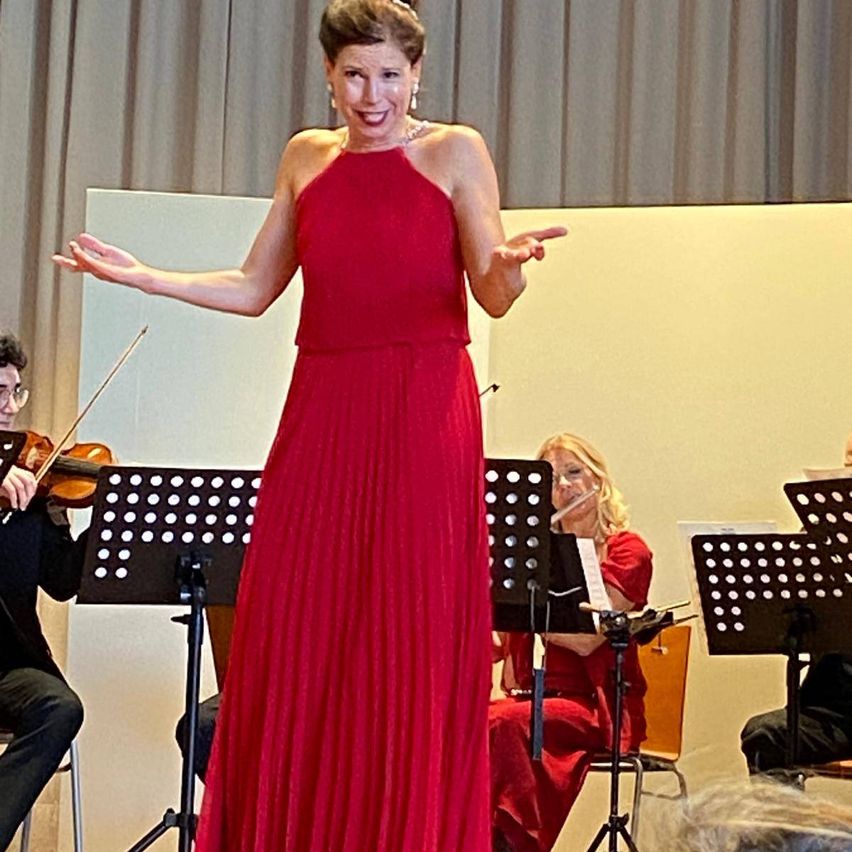 Eine Frau in einem roten Kleid steht lächelnd auf der Bühne, während ein Violinist und eine Flötistin im Hintergrund spielen.