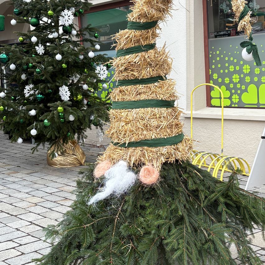 Ein Außenbereich mit einem Strohmann und einem geschmückten Weihnachtsbaum im Hintergrund. Der Mann hat einen Bart und trägt einen Hut. In der Nähe zeigt ein Geschäft eine festliche Fensterdekoration.