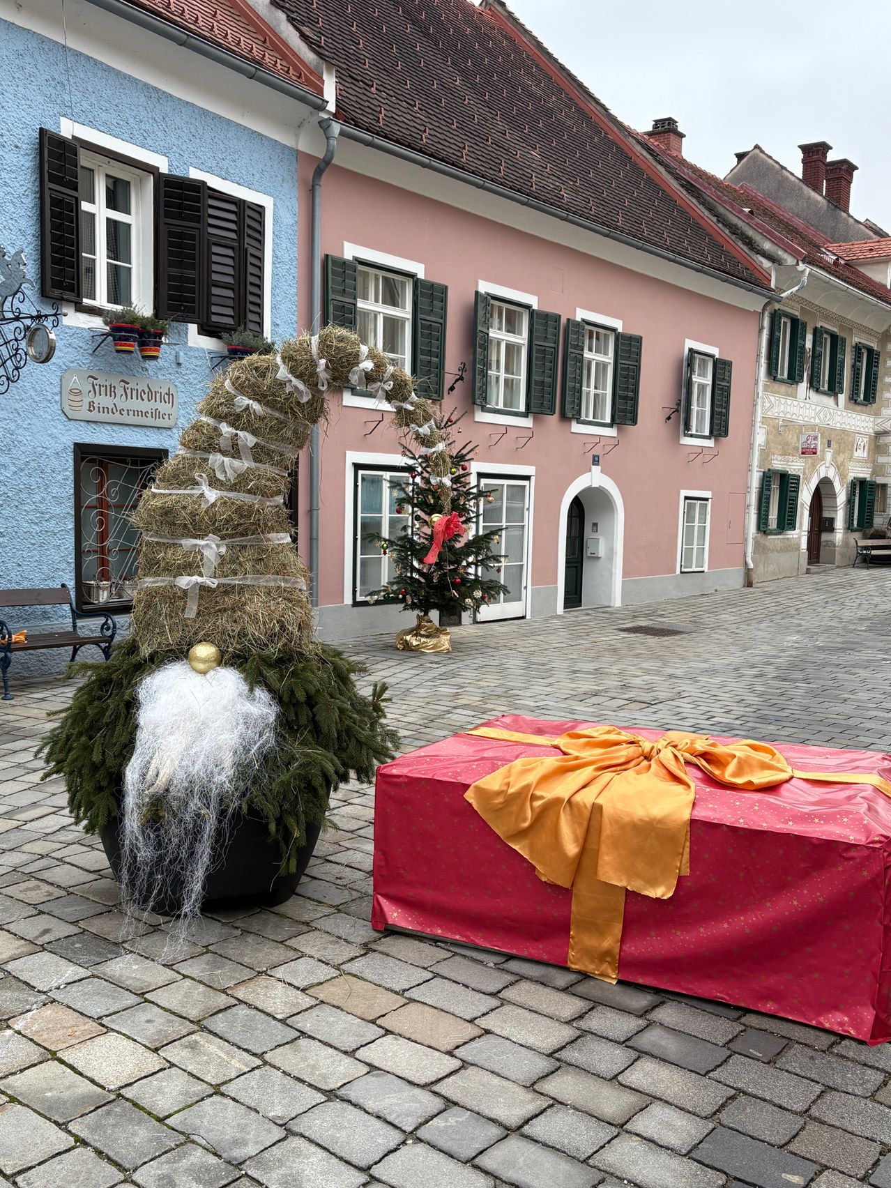 Eine zwergförmige Weihnachtsdekoration aus Stroh und Pflanzen steht neben einem roten Geschenkkarton mit gelber Schleife auf einer Kopfsteinpflasterstraße. Die Straße ist von bunten Gebäuden gesäumt, von denen eines einen Weihnachtsbaum vor sich hat.