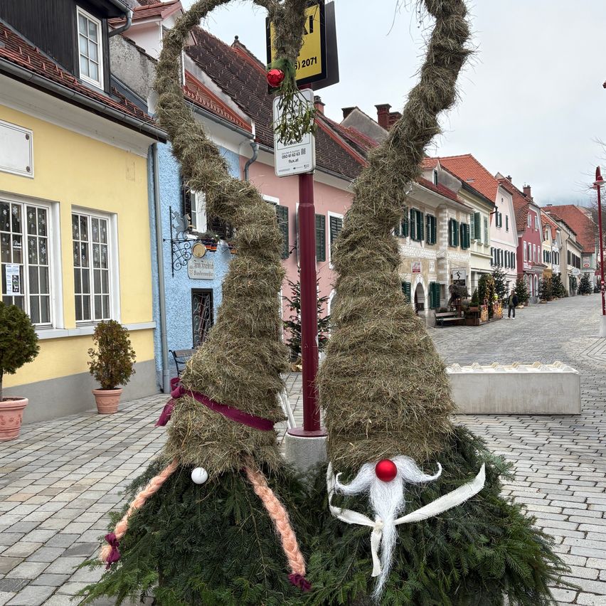 Zwei aus Stroh und Grün hergestellte Zwerge stehen auf einer Kopfsteinpflasterstraße, dekoriert für Weihnachten. Dahinter befinden sich bunte Gebäude, ein Schild mit der Nummer 2011 und eine Straßenlaterne.