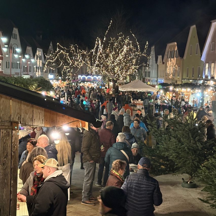 Ein überfüllter Outdoor-Markt bei Nacht mit festlichen Lichtern und Tannenbäumen. Die Leute kaufen an Ständen unter einem Zelt. Im Hintergrund sind Gebäude mit historischer Architektur zu sehen.