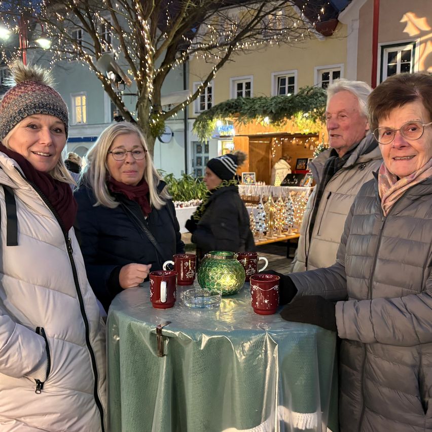 Vier Erwachsene stehen um einen Tisch auf einem Weihnachtsmarkt und lächeln für ein Foto. Hinter ihnen sind Geschäfte mit festlichen Lichtern geschmückt. Eine Frau mit Brille und Schal hält eine Tasse.