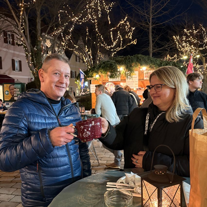 Ein Mann und eine Frau lächeln bei einer Veranstaltung im Freien, halten Tassen, mit einem Laternen und Tischsetting dahinter und beleuchteten Bäumen im Hintergrund.