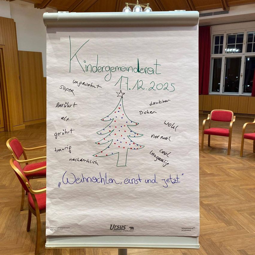 Ein Flipchart mit einer Zeichnung eines Weihnachtsbaums und Wörtern auf Deutsch, auf einem Holzboden platziert. Rote Stühle umgeben den Bereich.