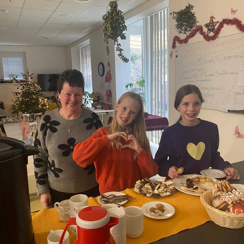 Eine Frau und zwei Mädchen posieren für ein Foto in einem Essbereich. Sie lächeln und zeigen ihre Hände. Vor ihnen steht ein Tisch mit Tellern mit Keksen, Tassen und einem Kaffeepott. Der Essbereich hat einen Weihnachtsbaum, eine Tafel und einige Dekorationen.