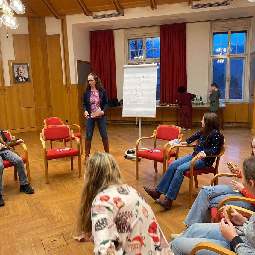 Eine Gruppe von Menschen sitzt in Stühlen um eine Frau, die steht und spricht. Es gibt eine Tafel, einen Tisch mit Flaschen und ein gerahmtes Bild an der Wand.