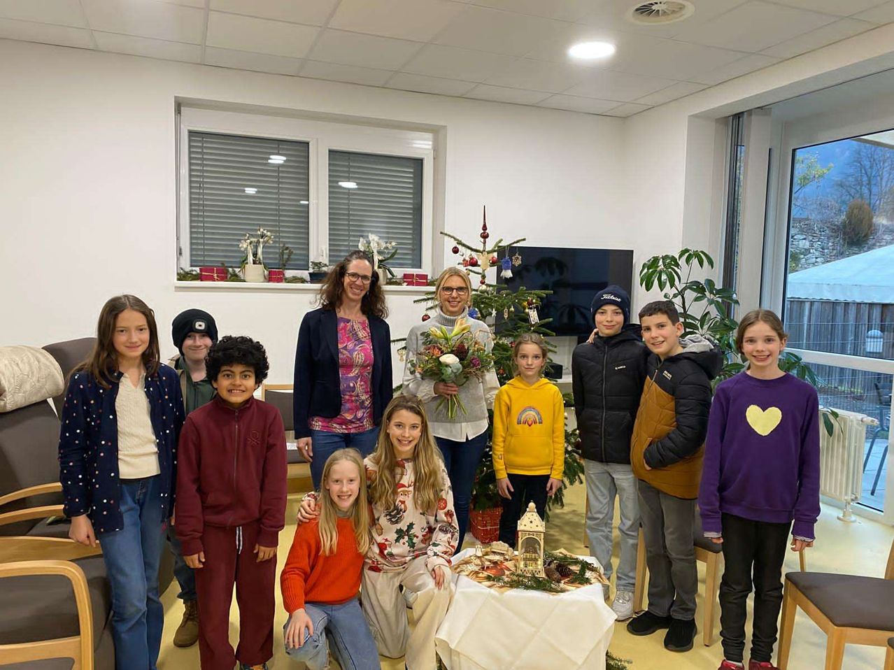 Eine Gruppe von Kindern und Erwachsenen posiert für ein Foto in einem Raum mit einem Weihnachtsbaum, einem Tisch mit Dekorationen und einem Fernseher an der Wand. Die Kinder tragen Winterkleidung und lächeln.