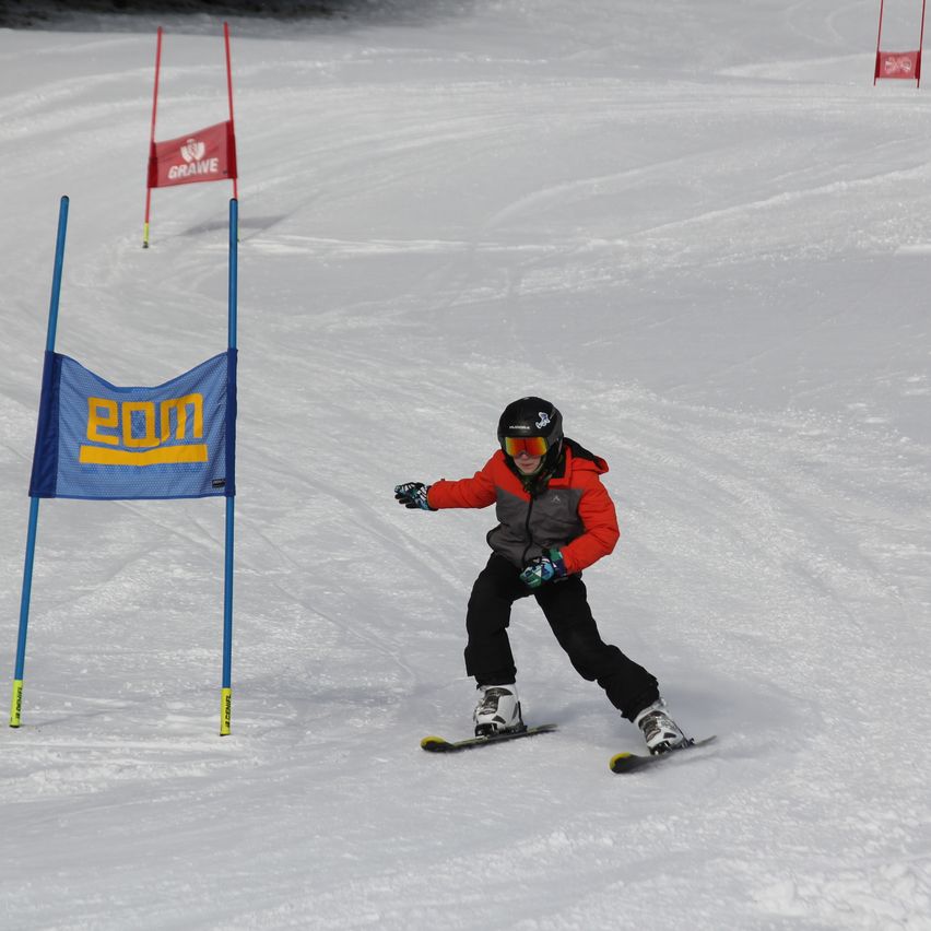 Ein Skifahrer navigiert einen schneebedeckten Hang mit blauen Pfosten und Schildern. Er trägt einen rot-grauen Mantel und schwarze Hosen.