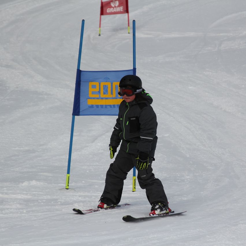 Ein Skifahrer in einem schwarzen Helm und Skianzug fährt den Hang hinunter. Hinter ihm steht ein blaues Schild mit gelben Buchstaben. Ein roter Flagge mit dem Wort 'GRAWE' ist in der Ferne.