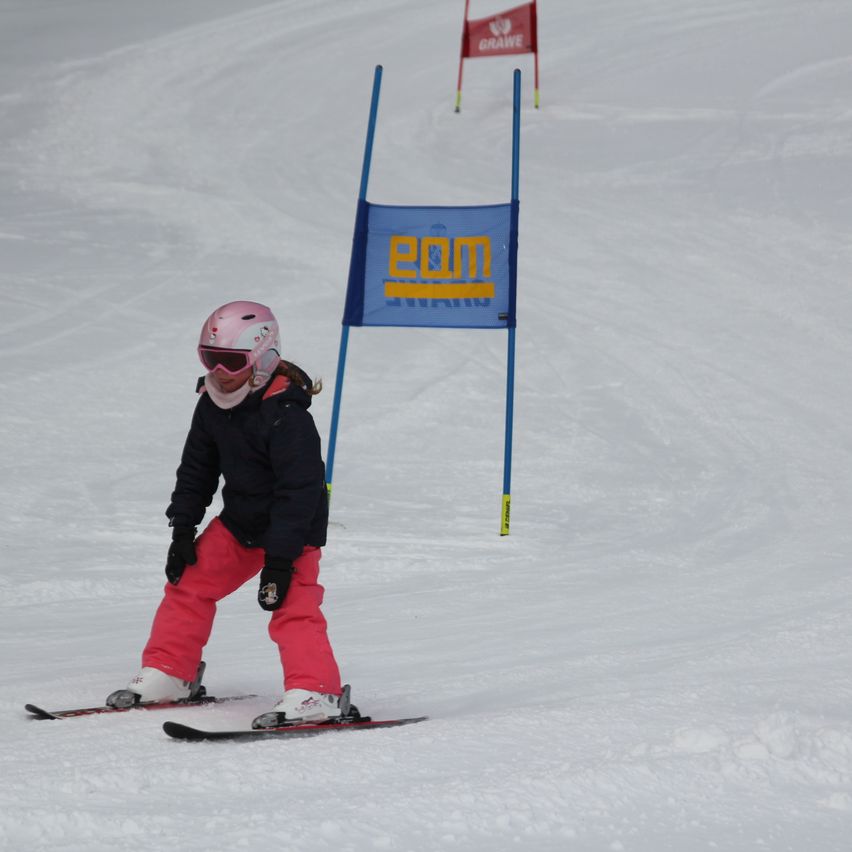 Ein junger Skifahrer mit einem rosa Helm fährt einen verschneiten Hang hinunter, wobei ein blaues Schild mit der Aufschrift 'pom' im Hintergrund zu sehen ist.