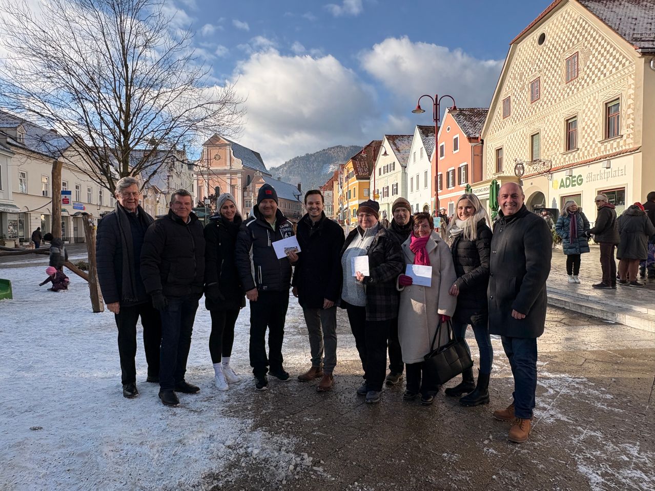 Eine Gruppe von Menschen steht auf einem verschneiten Stadtplatz, einige halten Papiere in den Händen, während ein Gebäude mit einem Glockenturm und einem Baum dahinter steht.