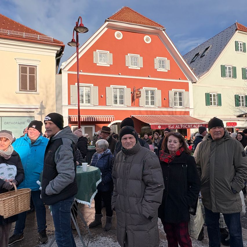 Eine Gruppe von Menschen, die warm gekleidet sind, steht vor Gebäuden. Eine Frau hält einen Korb, und ein Mann steht bei einem Tisch. Ein Gebäude mit roten Wänden und grünen Fenstern hat ein rotes Vordach.