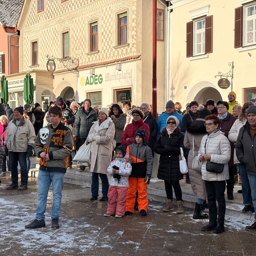 Eine Menschenmenge versammelt sich vor einem Gebäude mit dem Schild 'ADEG Marktplatz' davor. Sie sind in Winterkleidung gekleidet und einige tragen Mützen. Einige stehen, andere sitzen. Der Boden ist schneebedeckt.