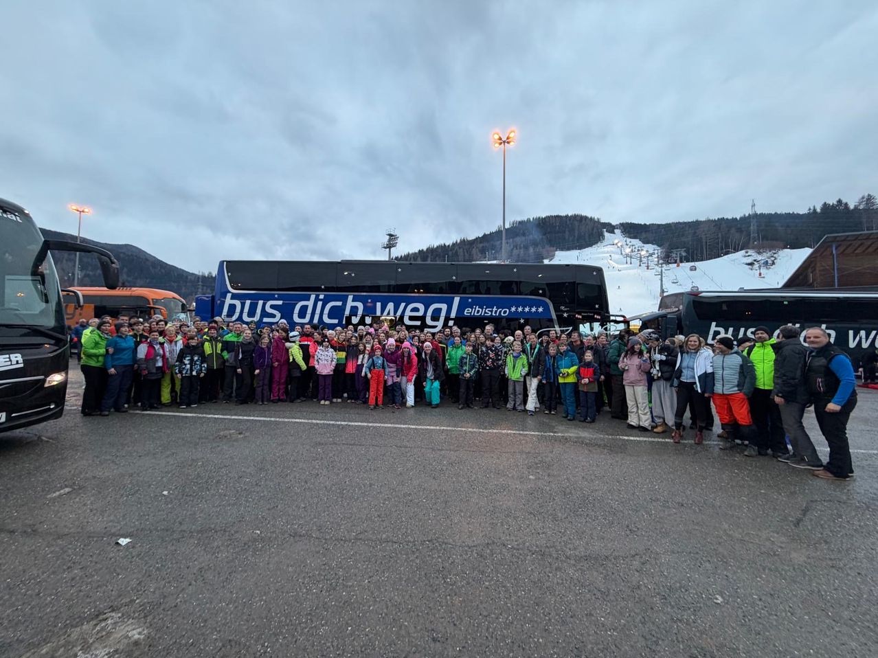 Eine Gruppe von Menschen posiert für ein Foto vor einem Bus mit dem Text 'bus dich weg! eibisstro' in einem verschneiten Gebiet.