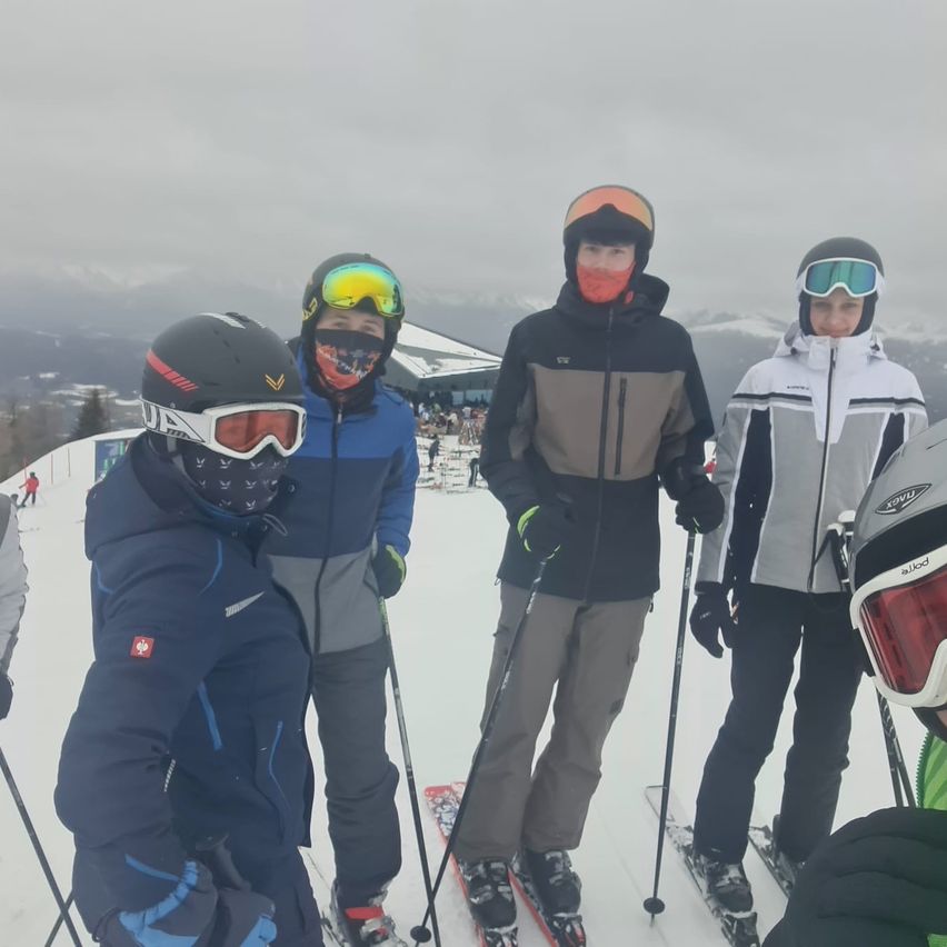 Fünf Skifahrer stehen auf einem verschneiten Berg, gekleidet in Winterausrüstung. Der Skifahrer in der Mitte trägt eine rote Gesichtsmaske. Hinter ihnen ist ein Gebäude zu sehen, und in der Ferne sind Menschen und Berge zu erkennen.