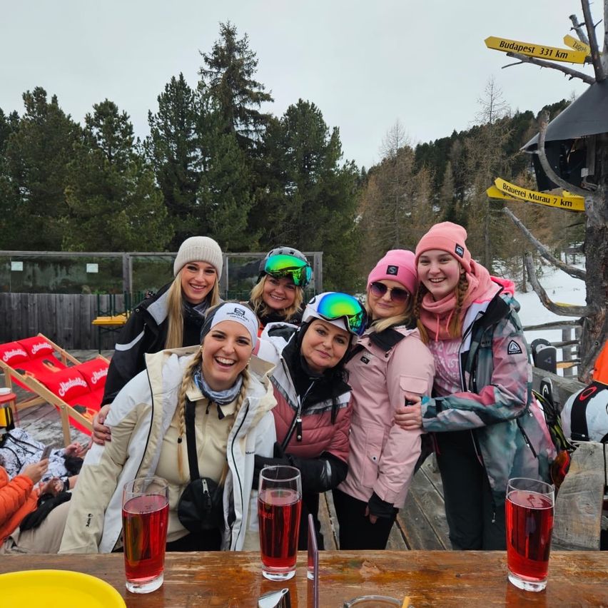 Eine Gruppe von Frauen posiert für ein Foto auf einem verschneiten Berg, trägt Winterkleidung und lächelt. Hinter ihnen stehen Bäume, ein Zaun und ein Schild. In der Nähe sitzen Leute auf Liegestühlen und ein Holztisch mit Gläsern roter Getränke.