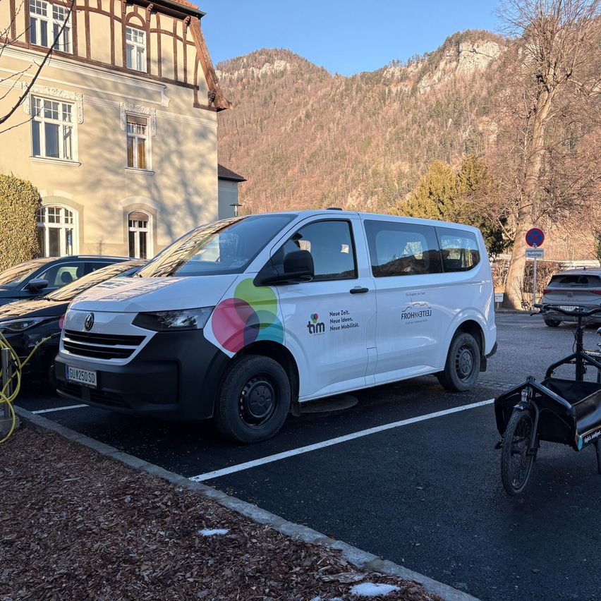 Ein weißer Van ist auf einem Parkplatz vor einem Gebäude mit Bergen im Hintergrund geparkt. Ein Fahrrad mit Anhänger steht in der Nähe.