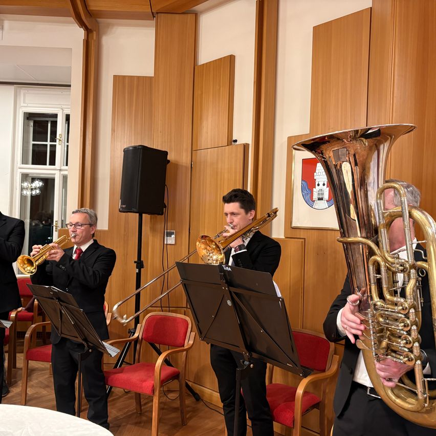 Eine Brass-Band in formellen Anzügen spielt Trompeten und Tubas in einem Raum mit Holzwänden. Die Musiker stehen vor Notenständern, mit roten Stühlen dahinter.