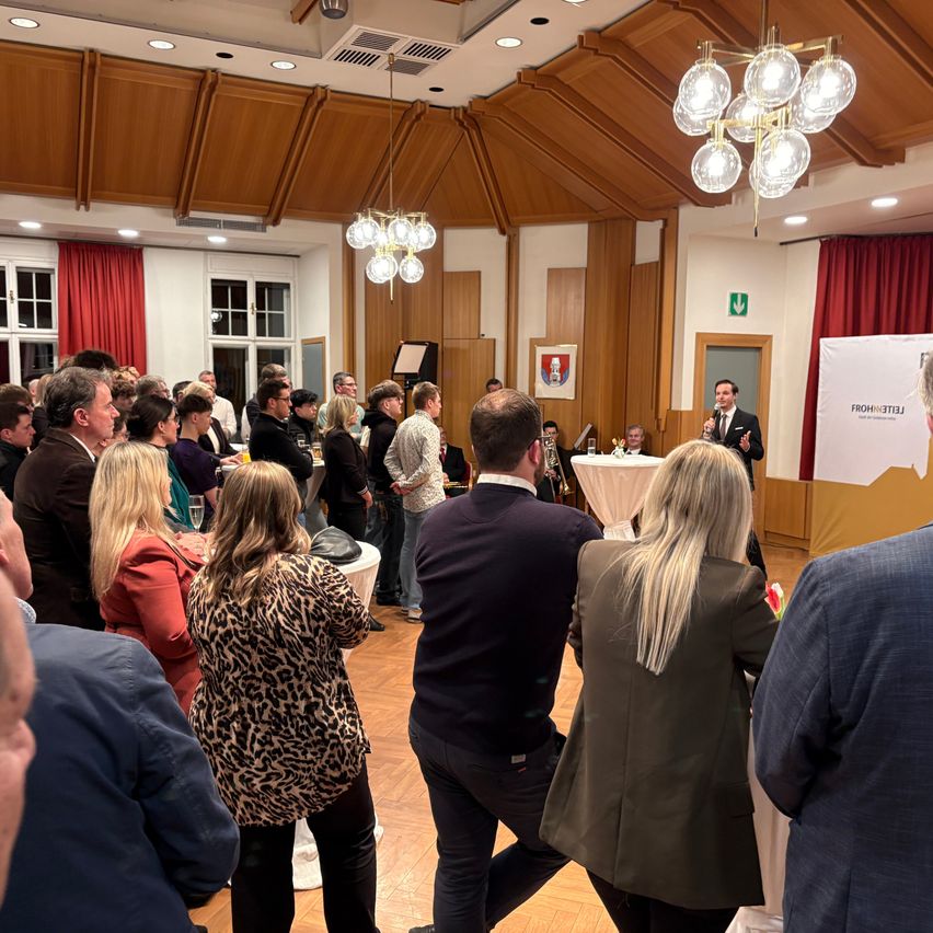 In einem Raum spricht ein Mann in ein Mikrofon. Dahinter steht ein Tisch mit weißer Tischdecke. Ein Banner mit Text steht auf der rechten Seite. Menschen stehen und lauschen.