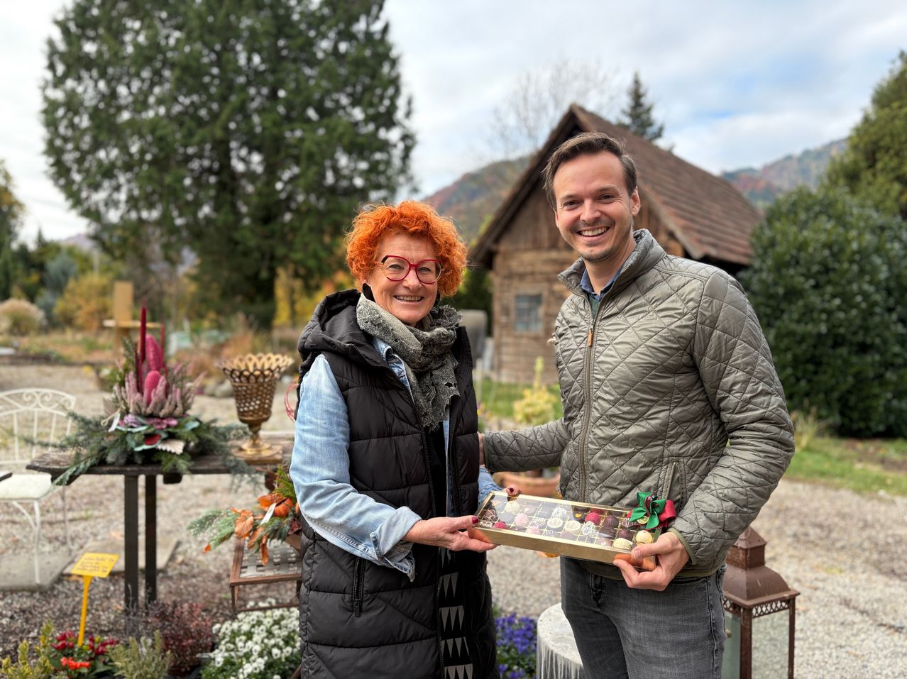 Eine Frau und ein Mann stehen lächelnd vor einem rustikalen Haus. Die Frau hält eine Schachtel Pralinen, während der Mann den Deckel hält. Sie sind von dekorativen Pflanzen und Blumen umgeben.