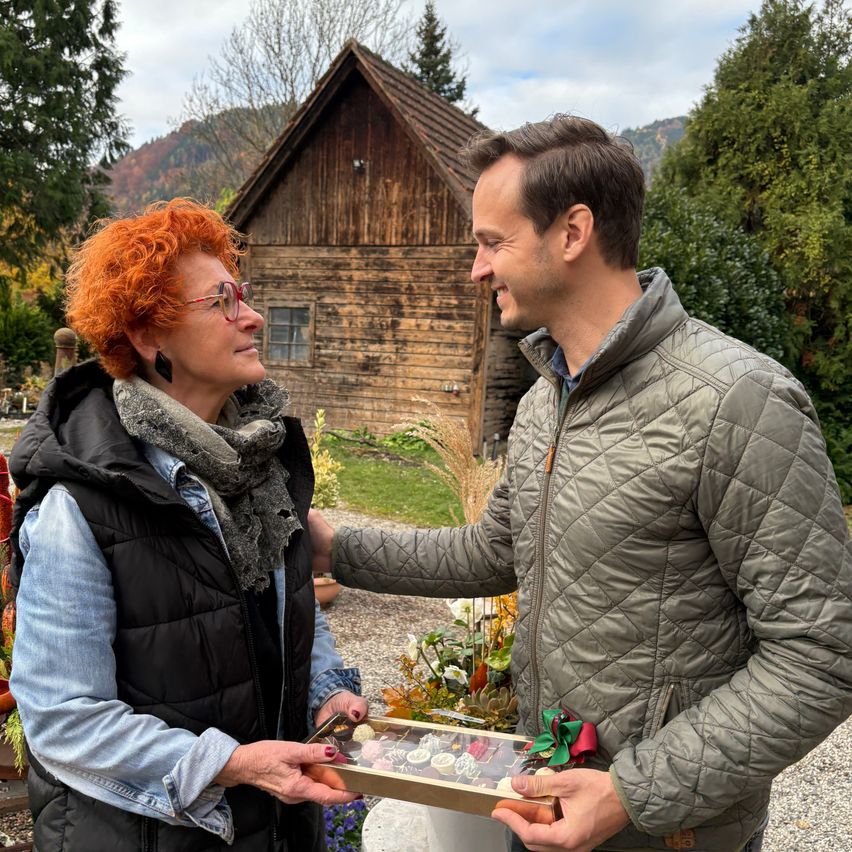 Ein Mann und eine Frau stehen vor einem Holzhaus, der Mann lächelt und hält eine Schachtel Schokolade. Die Frau hat rote Haare und trägt eine Brille.