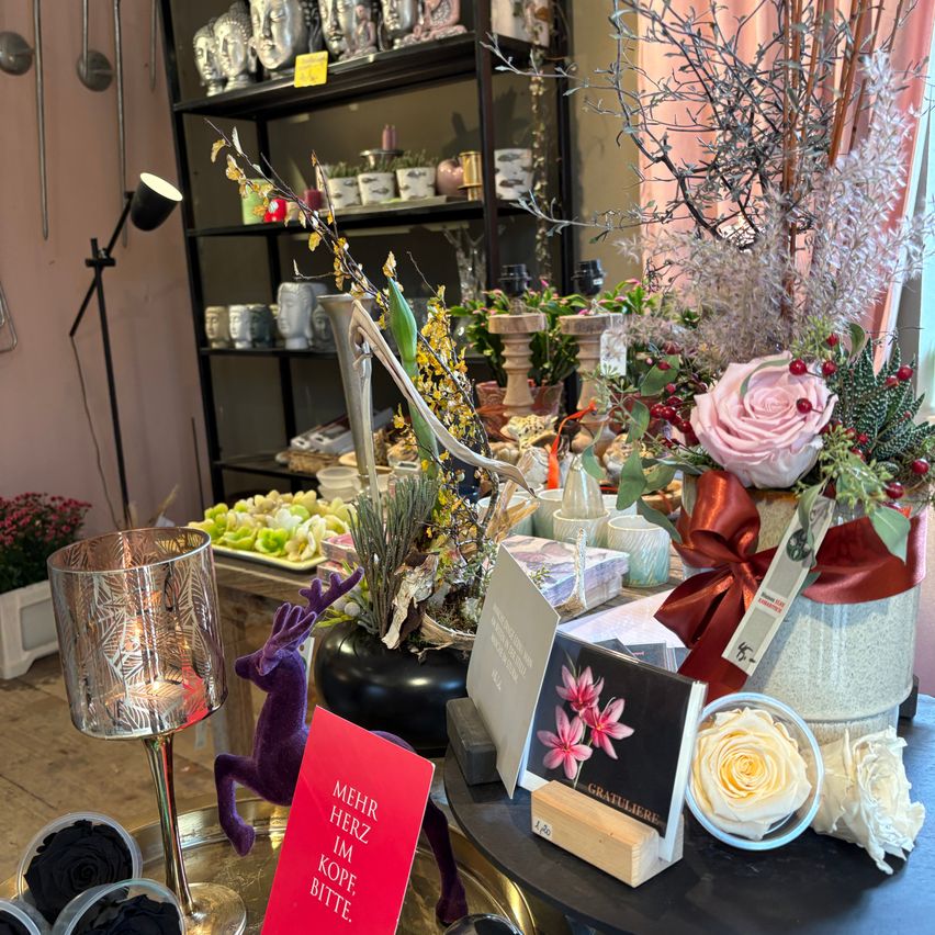 Ein Ladeninneres mit einem Tisch, der verschiedene Artikel präsentiert. Es gibt Blumenarrangements, dekorative Kerzen und eine Karte, die 'Mehr Herz im Kopf, bitte' lautet. Dahinter stehen Regale mit verschiedenen Artikeln wie Statuen und Tassen.