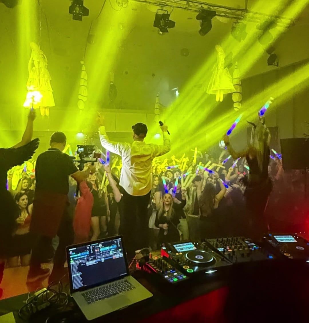 Ein DJ-Set in einem Club mit gelben Lichtern, einem DJ-Booth und einer tanzenden Menge mit Glühstäben. DJs auf der Bühne halten Mikrofone und treten auf.