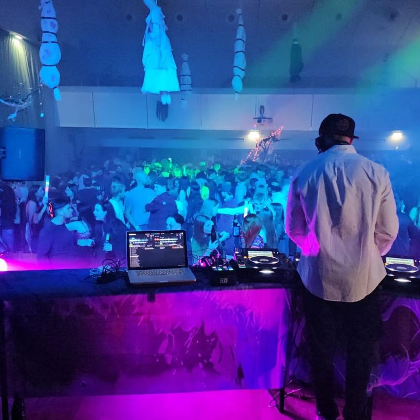 Ein Mann steht hinter einem Tisch mit Laptop und DJ-Equipment. Eine Menschenmenge tanzt in einem Raum mit blauen und lila Lichtern.