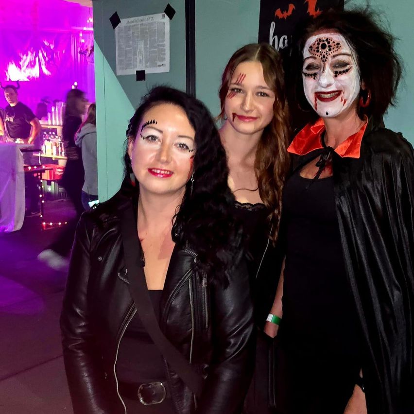 Drei Frauen stehen zusammen auf einer Party. Die Frau in der Mitte hat Make-up im Gesicht und die Frau rechts ist als Vampir verkleidet.