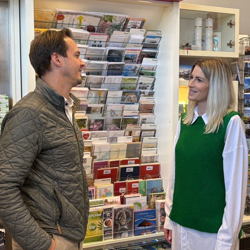 A man and a woman are standing in a shop, facing each other. The man is wearing a jacket, and the woman is wearing a white shirt and a green vest. Behind them is a display of cards.