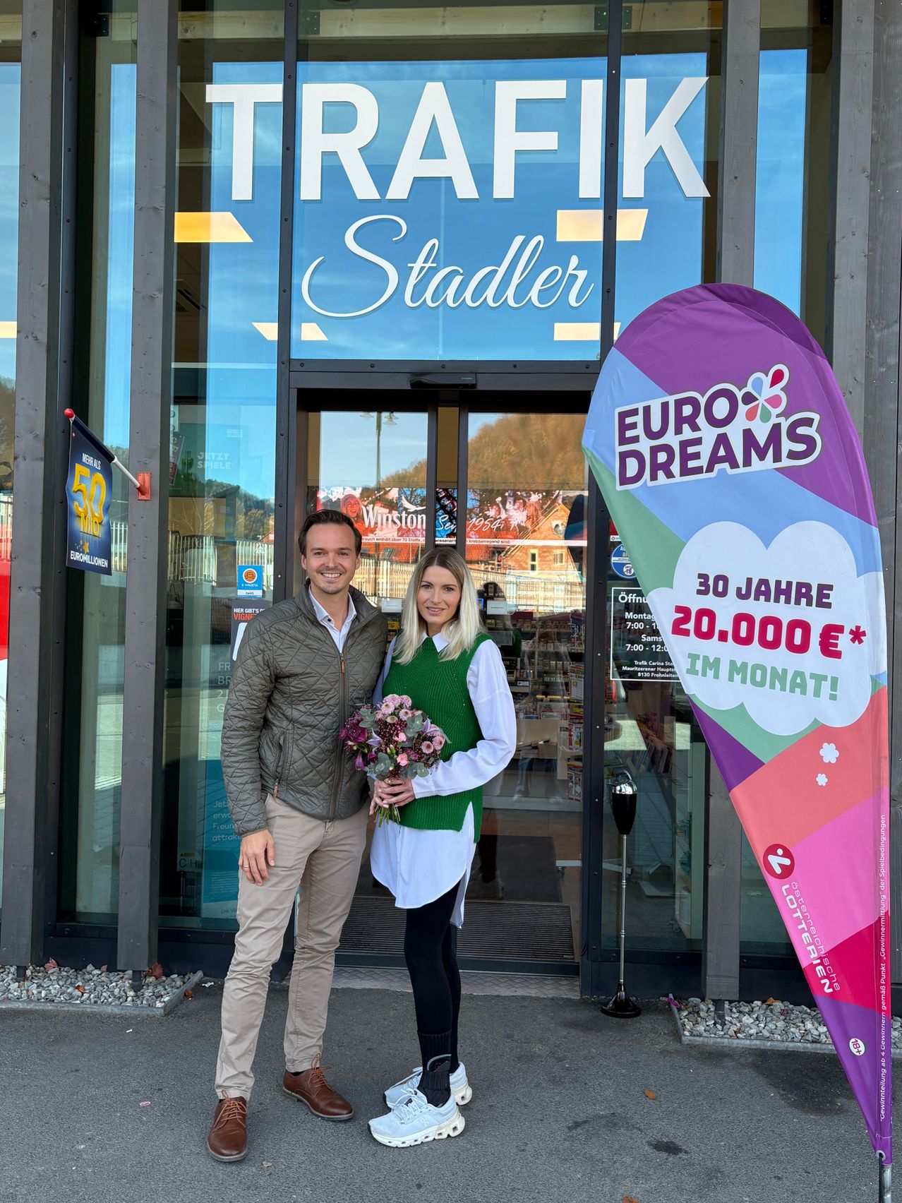 A man and woman stand in front of a store called Trafik Stadler, holding a bouquet of flowers. Behind them is a sign with Euro Dreams and a promotion for 30 years and 20,000 euros.