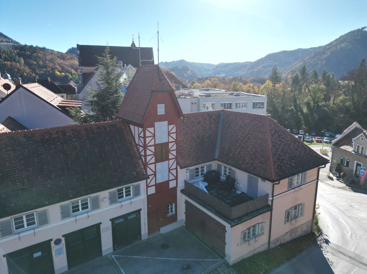 Aerial view of a quaint town with buildings, including a red-roofed house with white trim. The house features a balcony with a grill, and there are trees and mountains in the background.