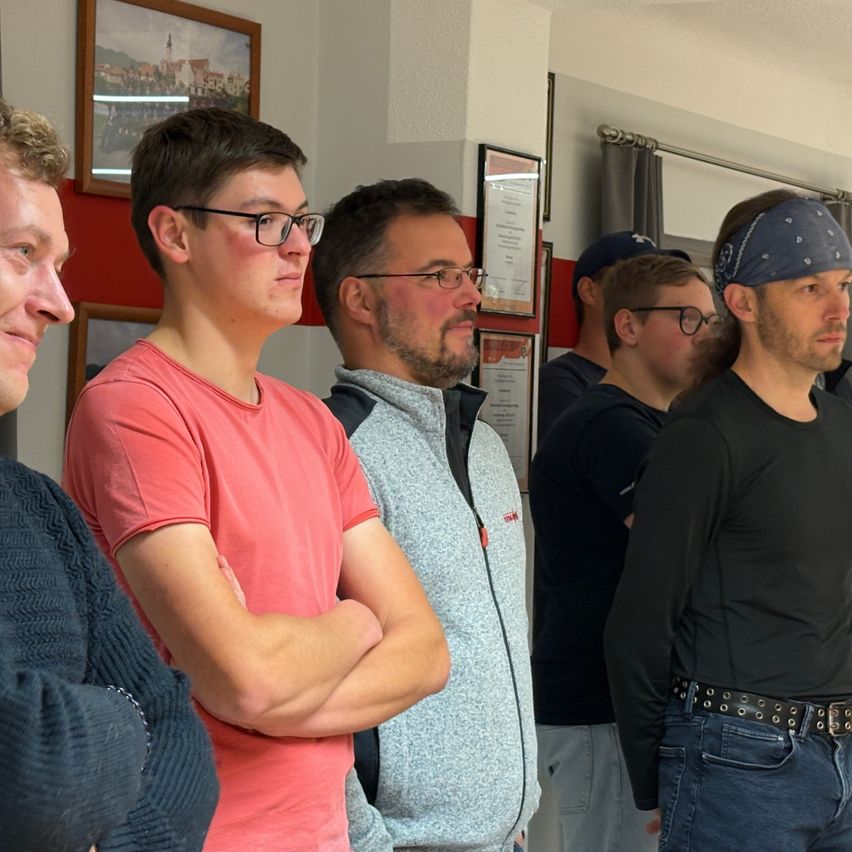 A group of men stands together in a room. One wears a bandana, another a belt. They appear focused, with one crossing his arms. Framed pictures adorn the wall.