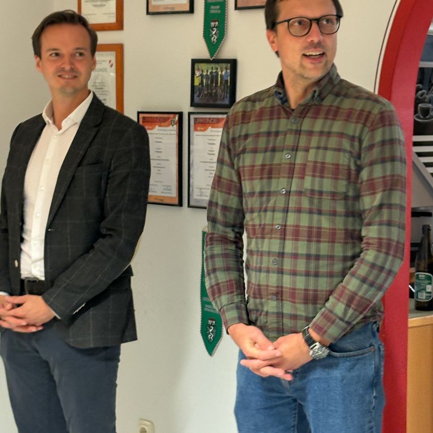 Two men stand in a room with a white wall, a green archway, and framed certificates. One wears a black jacket and white shirt, while the other wears a plaid shirt and jeans.