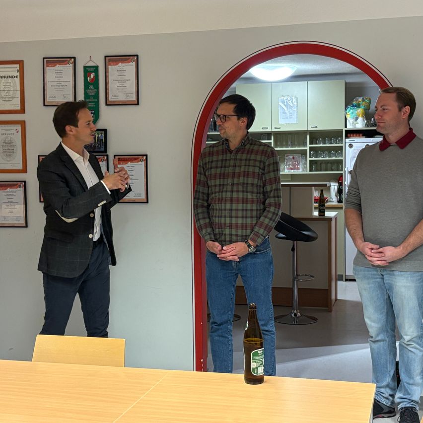 Three men stand in a room, two facing the camera, one behind a table. One man gestures, another wears glasses, and the third wears a red collar. Behind them, a framed certificate hangs on a wall.
