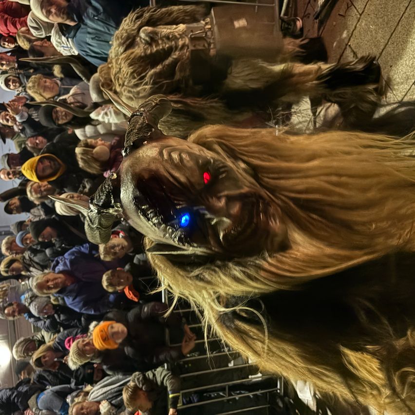 A crowd of people watches a large, hairy creature with glowing red eyes and a blue light on its head, standing in front of a barrier.