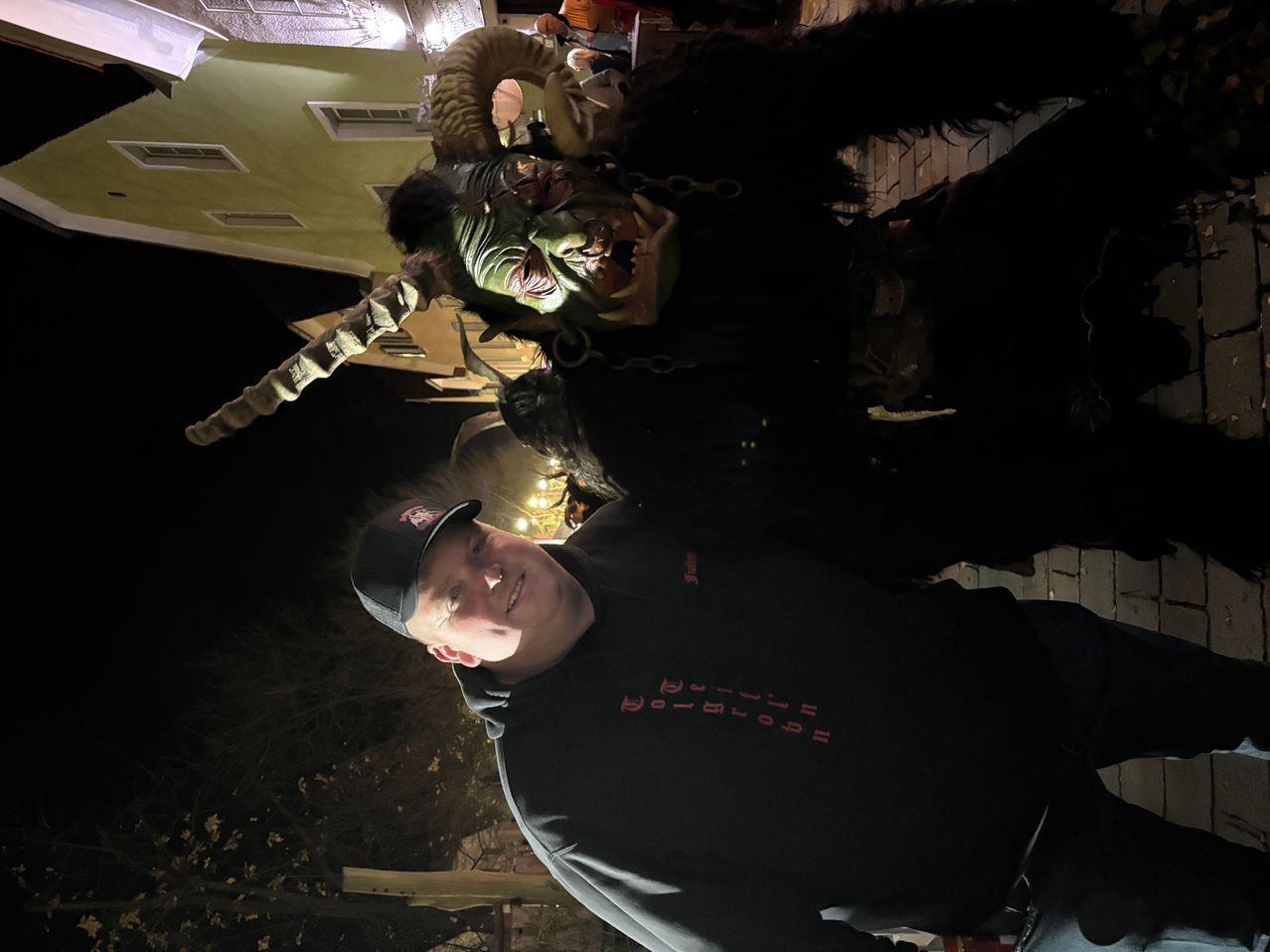 A man poses with a costumed character at night. The character wears a horned mask, black fur, and chains. The man is in a black sweatshirt with red text.
