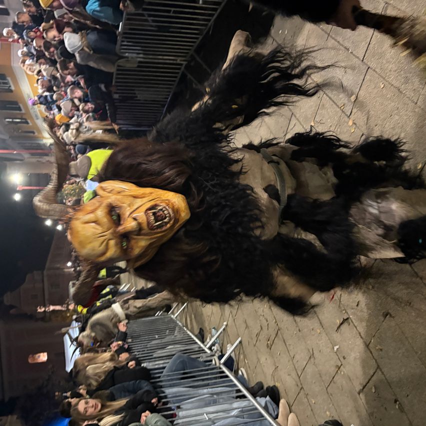 A crowd gathers at night to watch a person in a furry costume with a scary mask and horns crawling on the pavement.