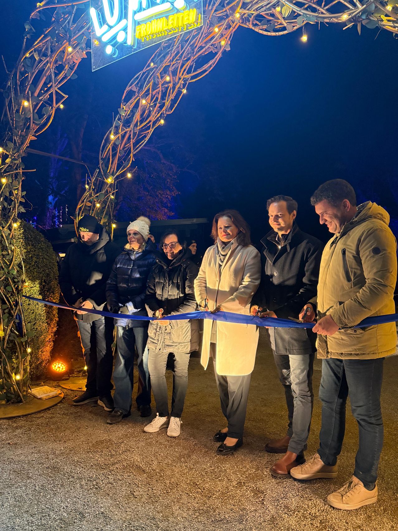 A group of people are standing together, smiling, and holding scissors. They are cutting a blue ribbon as part of a celebration or event. They are surrounded by trees and bushes with lights.