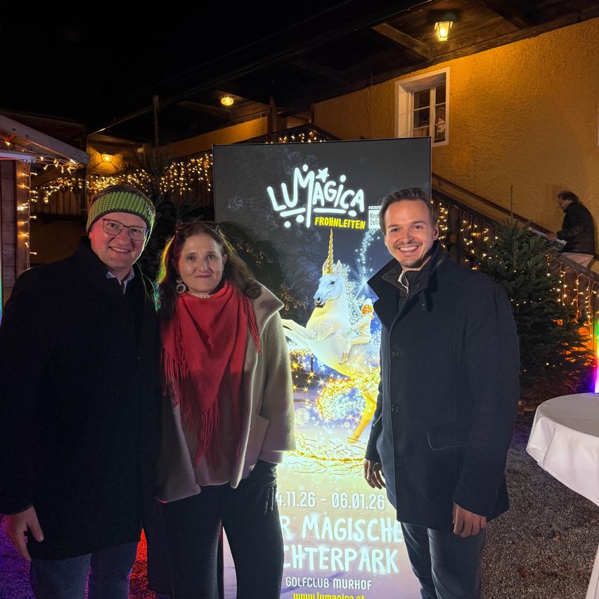 Three individuals stand next to a large sign for Lumina Magica, an event happening from November 11, 2026, to January 6, 2026. They are smiling and dressed for winter. Behind them, there are stairs, a Christmas tree, and a building with a window.