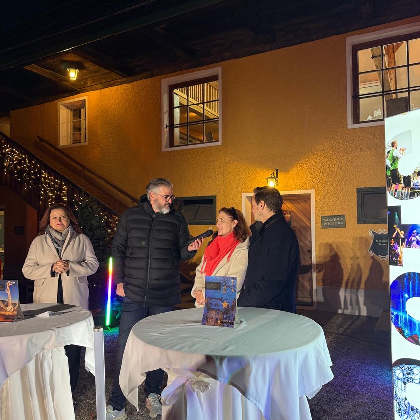 A man with a microphone speaks to a woman in a red scarf and a man in a black jacket at an event. They stand near a table with a book. Behind them is a building with windows and stairs.