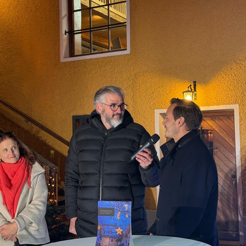 A man with a microphone speaks into the hand of another man, while a woman stands beside them. They are in front of a table with a book. Behind them is a wooden door with a lamp.