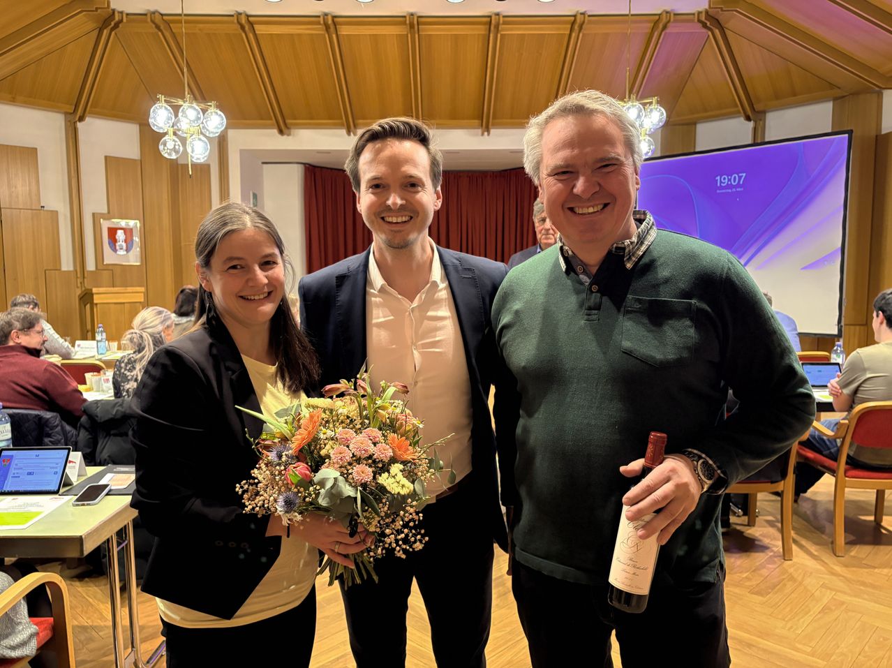 Drei Menschen stehen lächelnd in einem Raum mit Holzwänden und einer Bühne. Eine Frau hält Blumen, ein Mann hält eine Flasche und ein anderer Mann steht dazwischen.