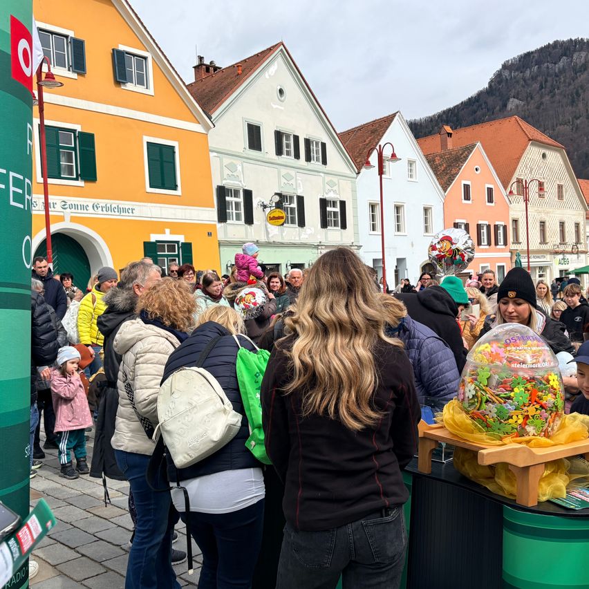 Eine Menschenmenge steht vor einem Gebäude mit vielen bunten Geschäften. Eine Frau mit blonden Haaren schaut sich ein großes Bonbon-Ei auf einem Holzständer an.