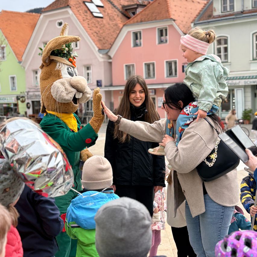 Ein Maskottchen verteilt Süßigkeiten an eine Gruppe von Kindern auf einem Stadtplatz, umgeben von Gebäuden und anderen Menschen.